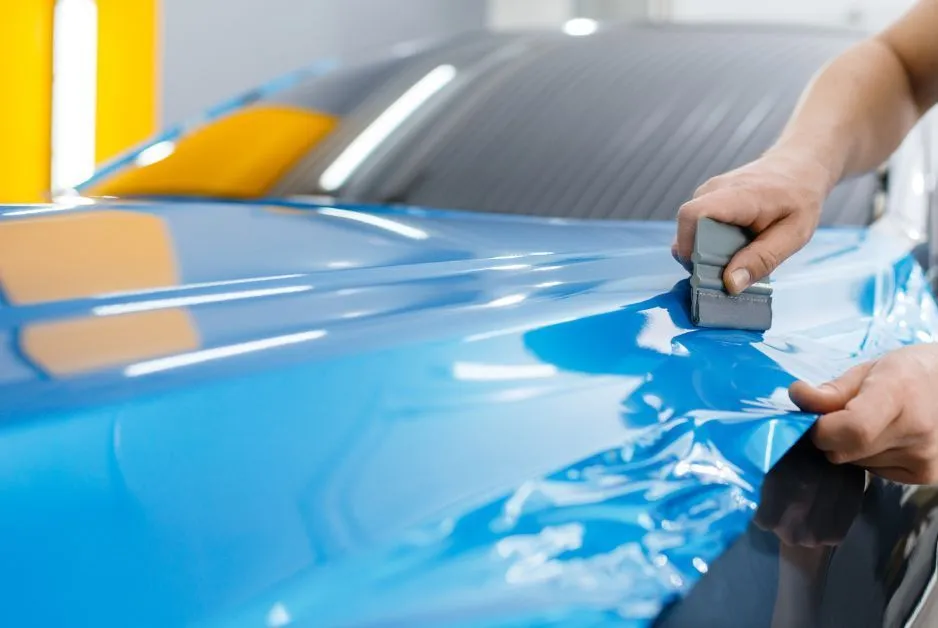 Person applying blue vinyl wrap to a car hood using a squeegee.