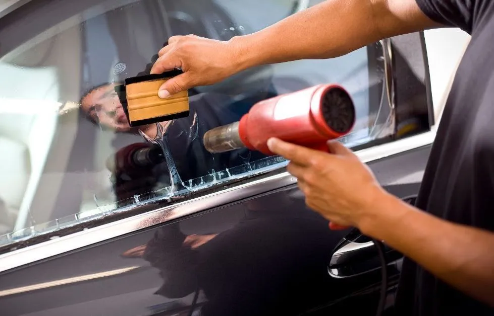 Person applying tint film to a car window with a heat gun and a squeegee.