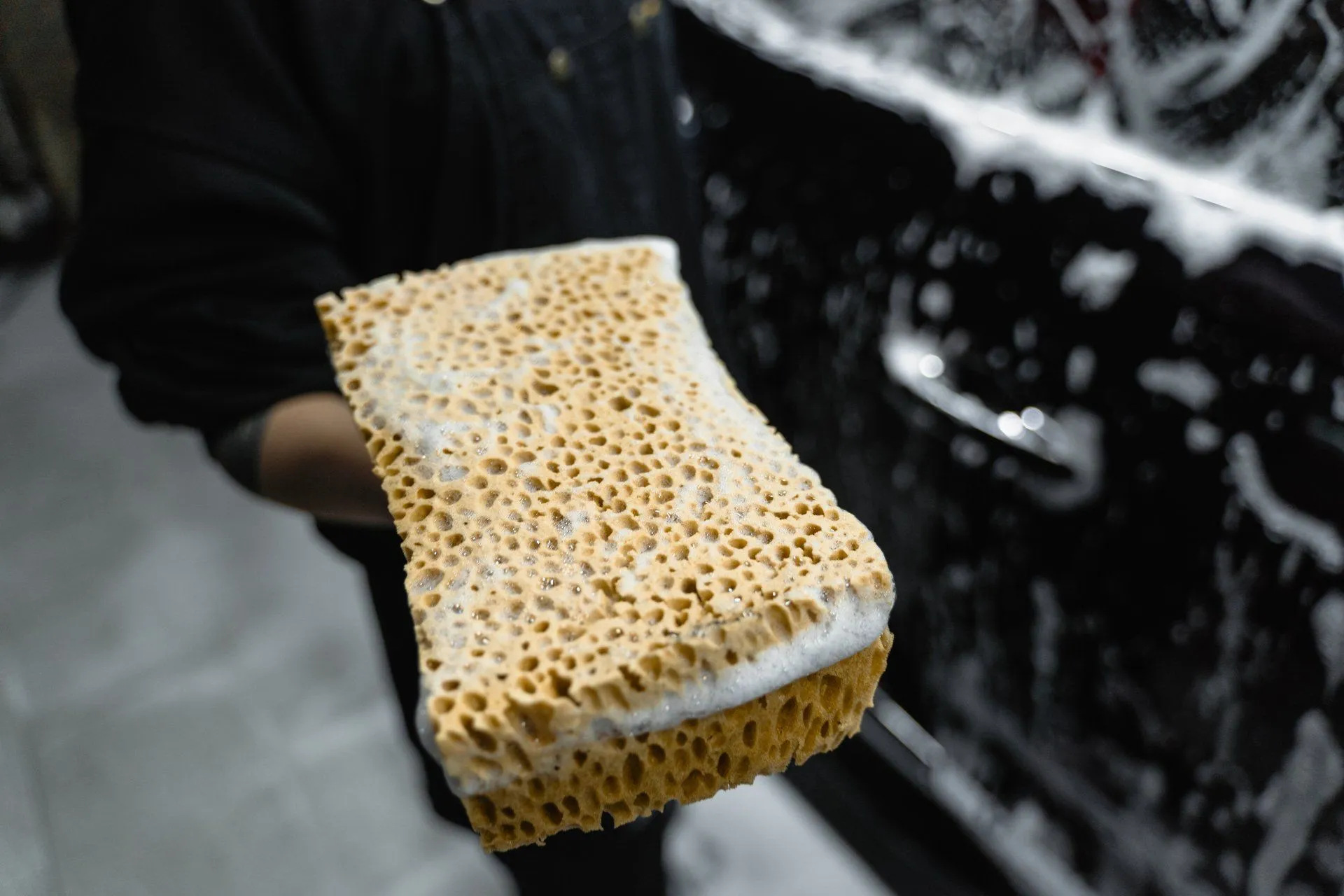 Person holding a wet, soapy sponge next to a black car door, washing the vehicle.